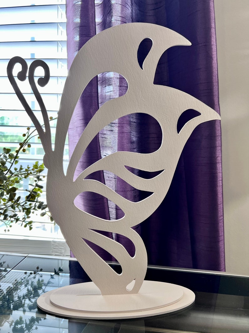 May include: White wooden butterfly sculpture with intricate cutouts. The butterfly is standing on a round base.
