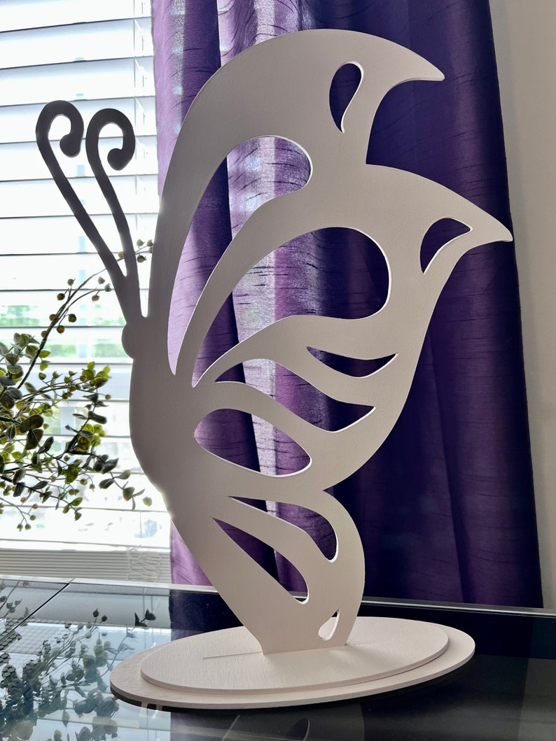 May include: White wooden butterfly cutout with a decorative base. The butterfly has a detailed design with cutouts in the wings. The base is a simple oval shape.