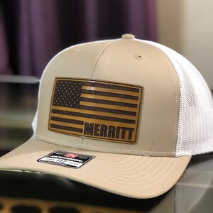 May include: A tan and white trucker hat with a brown leather American flag patch and the name "MERRITT" on it.
