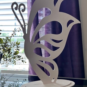 May include: White butterfly-shaped decorative wall art with a cutout design. The butterfly is standing on a white base.