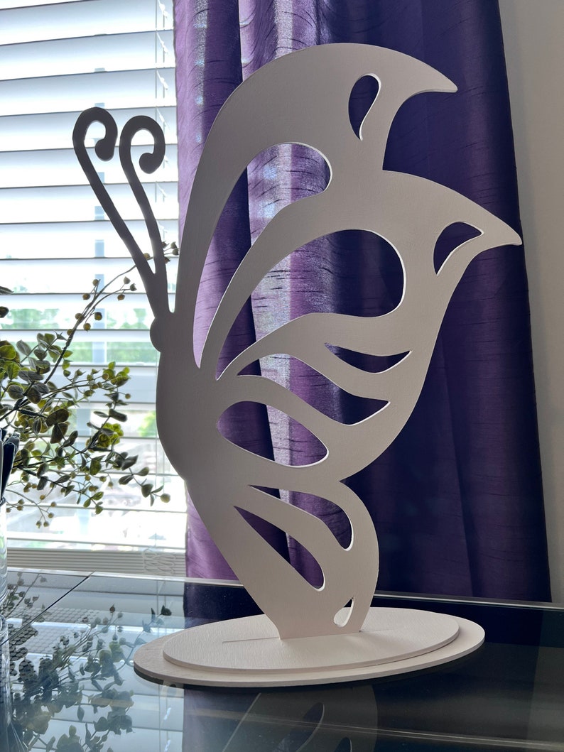 May include: White butterfly-shaped decorative wall art with a cutout design. The butterfly is standing on a white base.