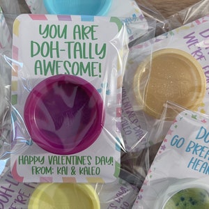 May include: Valentine's Day gifts featuring colorful playdough in clear packaging. Each package has a card with a playful message like "You are Doh-tally Awesome!" and "Happy Valentine's Day!" from Kai & Kaleo.
