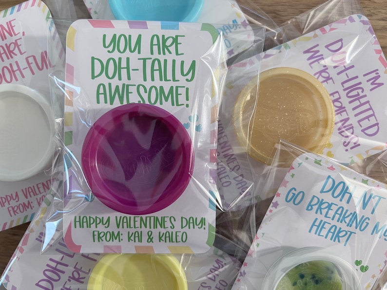 May include: Valentine's Day gifts featuring colorful playdough in clear packaging. Each package has a card with a playful message like "You are Doh-tally Awesome!" and "Happy Valentine's Day!" from Kai & Kaleo.