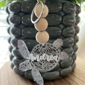 May include: A clear acrylic turtle keychain with the name "Amelia" engraved on the shell. The keychain has a silver metal ring and a string of wooden beads.