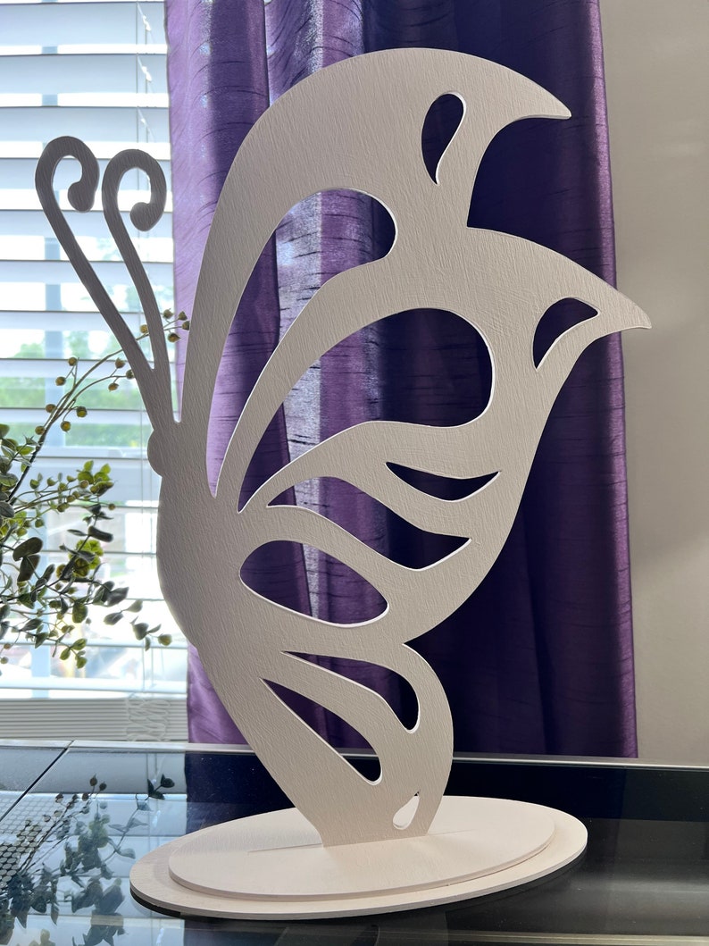 May include: White wooden butterfly sculpture with intricate cutouts. The butterfly is standing on a round base.