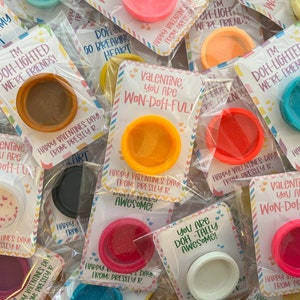 May include: Valentine's Day gifts featuring small, colorful containers of playdough. Each container is packaged with a card that has a Valentine's Day message. The colors of the playdough include pink, orange, blue, and white.