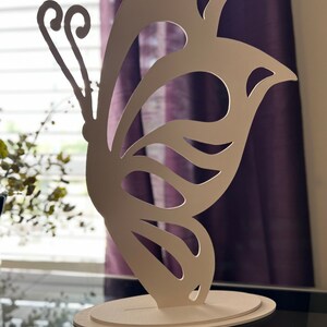 May include: White wooden butterfly sculpture with intricate cutouts. The butterfly is standing on a white oval base.