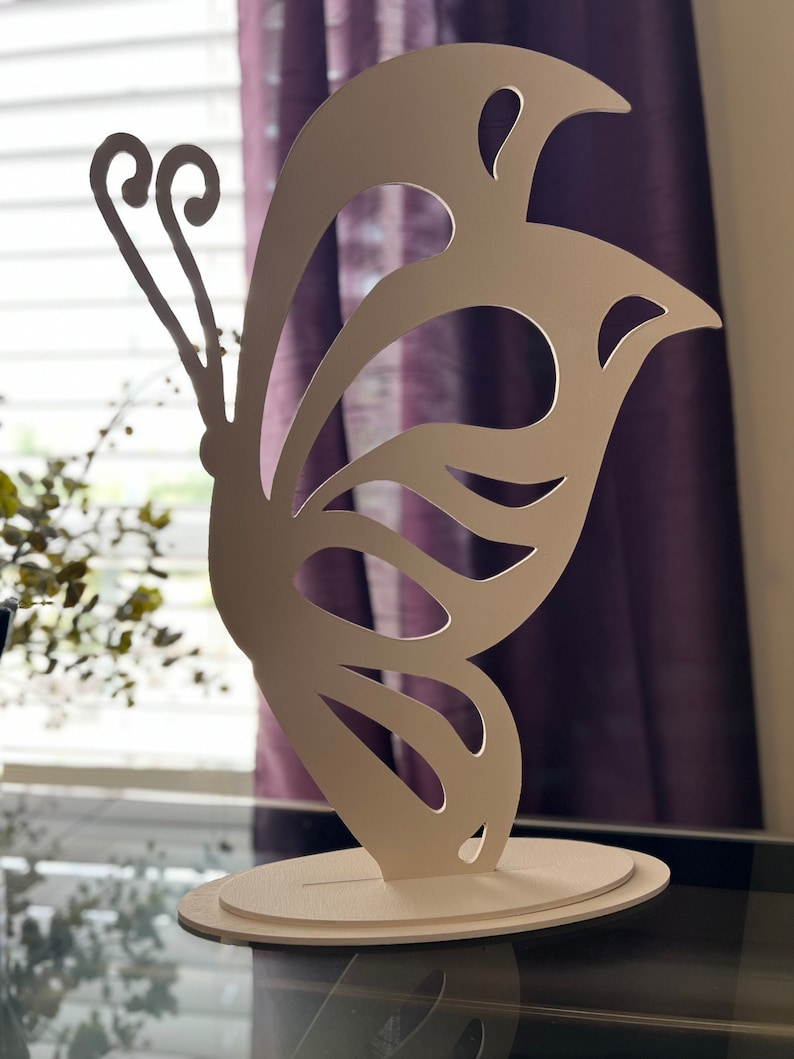 May include: White wooden butterfly sculpture with intricate cutouts. The butterfly is standing on a white oval base.