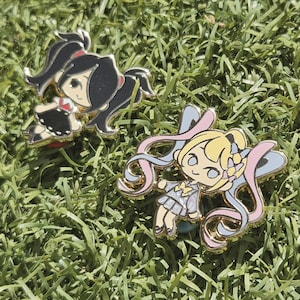 以下が含まれることがあります： 金色の縁取りが施された、アニメスタイルのキャラクターが描かれた2つのエナメルピン。1つのピンには、黒と赤のドレスを着た黒髪のキャラクターがいます。もう1つのピンには、ピンクと青のリボンが付いた、水色と銀色のドレスを着た金髪のキャラクターがいます。