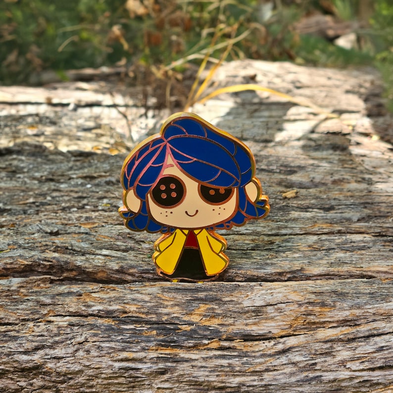 May include: A colorful enamel pin featuring a cartoon character with blue hair, button eyes, and a yellow coat. The pin has a gold outline and is set against a natural wood background.