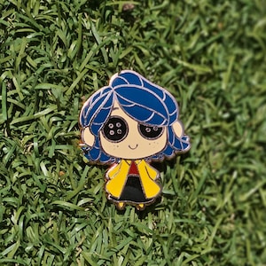 May include: Enamel pin featuring a cartoon character with blue hair, button eyes, and a yellow coat over a black dress. The pin has a gold outline and is set against a backdrop of green grass.