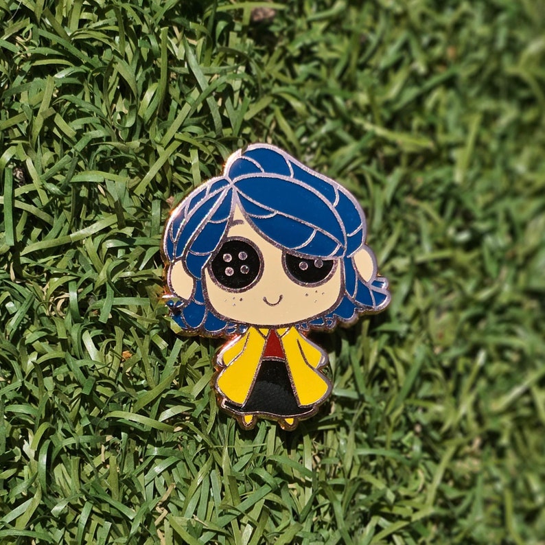 May include: Enamel pin featuring a cartoon character with blue hair, button eyes, and a yellow coat over a black dress. The pin has a gold outline and is set against a backdrop of green grass.