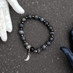 May include: A stretch bracelet featuring round grey mottled beads, small metallic spacer beads, a single smooth black bead, and a silver-toned charm shaped like a crescent moon. The bracelet is positioned between two decorative hand-shaped sculptures.
