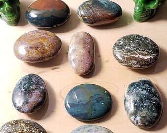 May include: A collection of ten polished stones, including a variety of colours and patterns, including green, brown, white, and grey. The stones are arranged in a circle on a wooden surface. Two green jade skull figurines are also included in the arrangement.