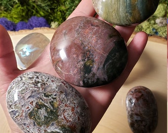 May include: A collection of six polished, smooth, oval-shaped stones with various patterns of green, brown, and white. The stones are arranged in a semi-circle on a wooden surface.