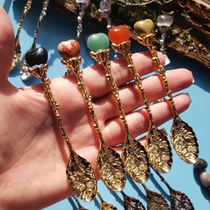 May include: Set of six gold-tone spoons with decorative leaf designs. Each spoon has a different coloured gemstone heart at the top of the handle. The gemstones are black, red, brown, green, orange, and yellow.