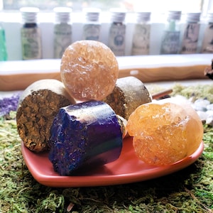 May include: A selection of raw crystals and stones in diverse forms and hues, including a deep blue, a golden orange, and a clear, spherical crystal. The crystals are displayed on a small, square, pink dish, set amidst green moss.