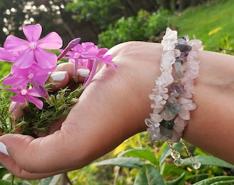 May include: A multi-strand bracelet made of small, irregularly shaped gemstones in shades of clear, pink, purple, and green. The bracelet is worn on a wrist, with a background of green foliage and pink flowers.