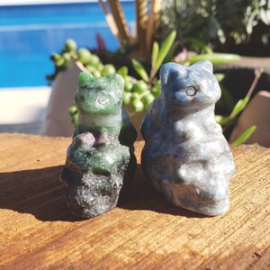 May include: Two carved stone squirrel figurines. One is green with red and black accents, the other is blue-grey. They are displayed on a wooden surface, with a blurred background of greenery and a pool.