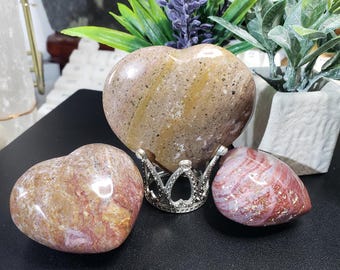 May include: Three heart-shaped polished stones, two are pink and red with white streaks, and one is brown with white and yellow streaks. The brown heart is on a silver stand.