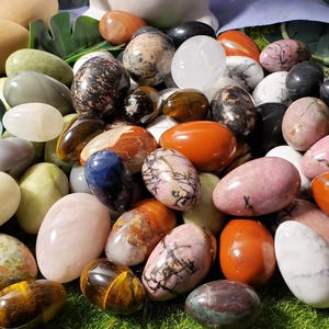 May include: A collection of polished, egg-shaped stones in various colors and patterns, including pink, red, green, white, and brown. The stones are arranged on a bed of green moss.