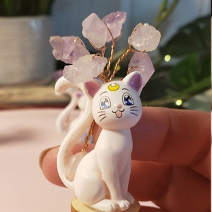 May include: A white ceramic figurine of a cat with a crescent moon on its forehead, sitting on a wooden base. The cat has blue eyes and a pink nose. A wire tree with purple amethyst crystals is attached to the cat's head.