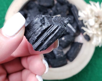 Peut inclure: Gros plan sur un cristal de tourmaline noire, montrant sa texture striée. Le cristal est tenu dans une main, avec d'autres cristaux de tourmaline noire visibles en arrière-plan.