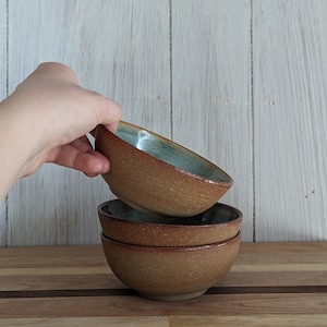 May include: Three brown ceramic bowls stacked on top of each other. The top bowl has a light blue glaze.