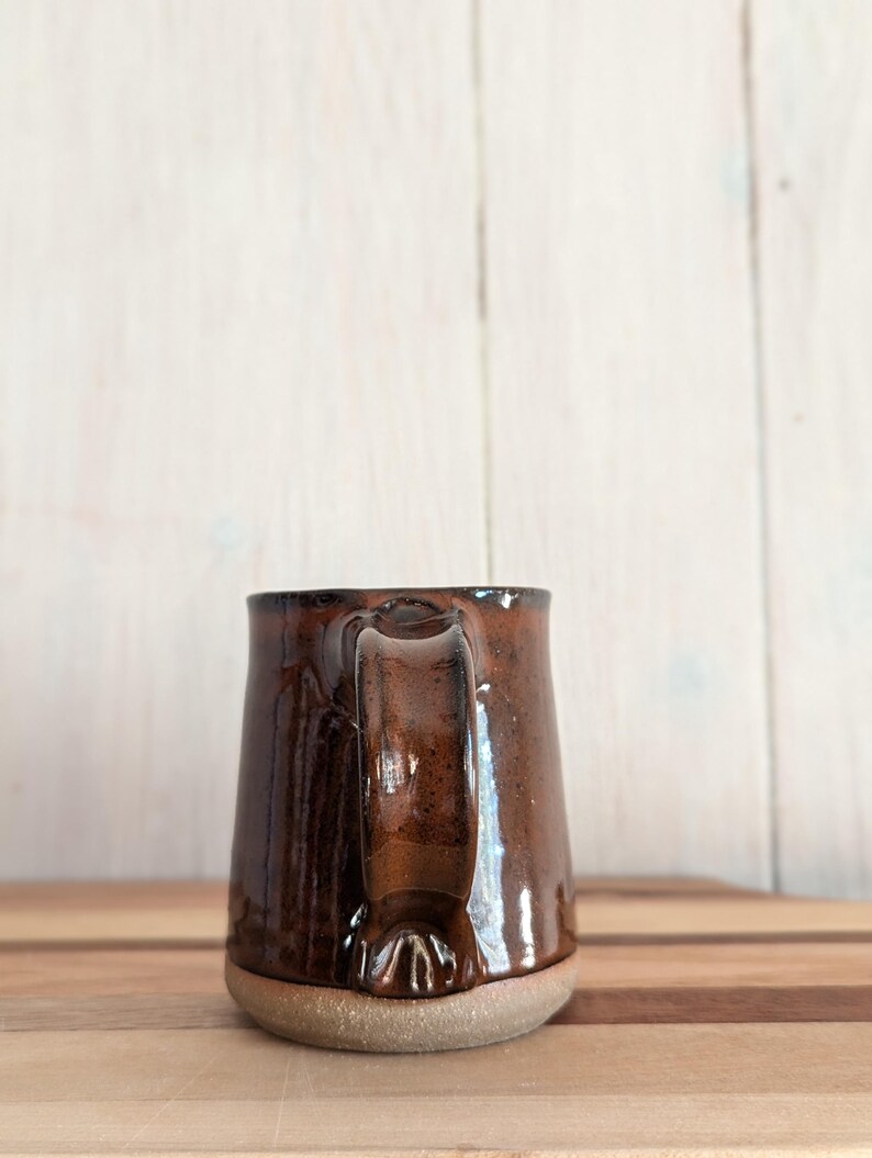 Puede incluir: Una taza de cer&aacute;mica marr&oacute;n con cuerpo redondeado y un asa ancha. La taza tiene un acabado brillante y una base ligeramente texturizada.