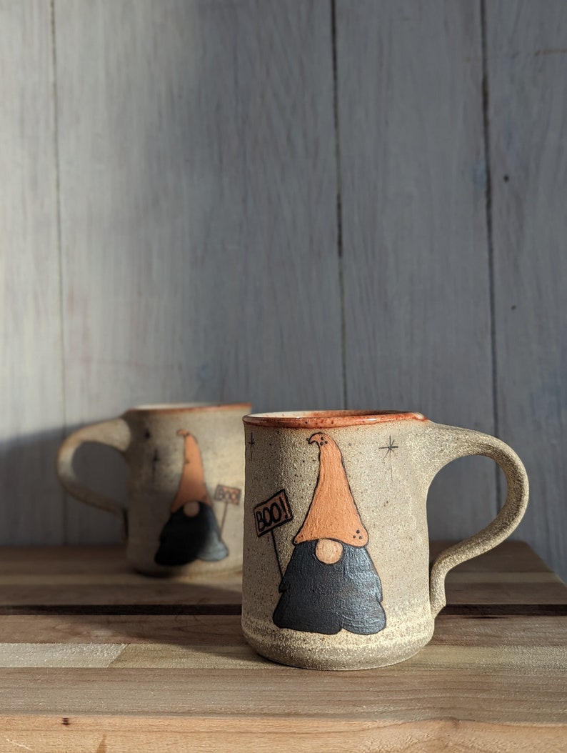 Stoneware mug with hand-painted halloween gnome with an orange hat and big brown beard with a round nose sticking out. The gnome is holding a sign that says "BOO!" and is surrounded by black stars.
