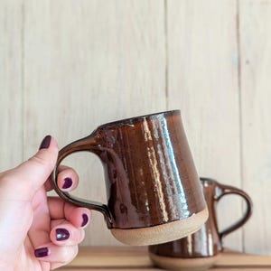 Puede incluir: Dos tazas de cer&aacute;mica marr&oacute;n con un esmalte moteado. La taza en primer plano est&aacute; siendo sostenida por una mano con esmalte de u&ntilde;as oscuro.