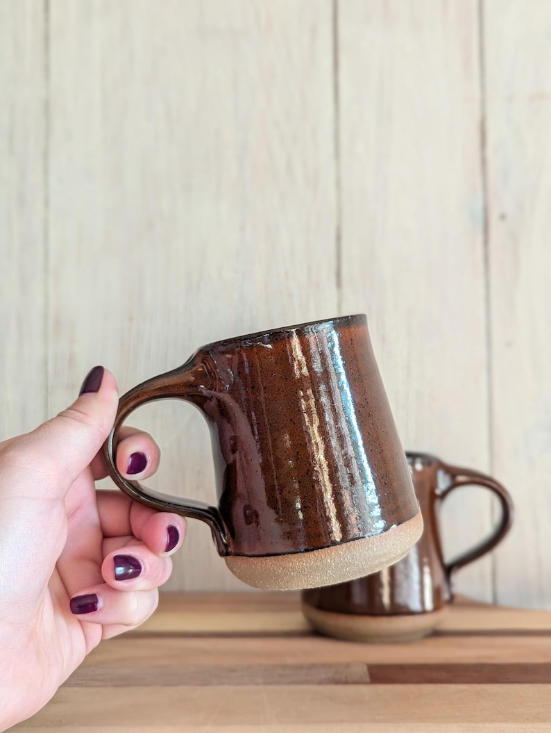 Puede incluir: Dos tazas de cer&aacute;mica marr&oacute;n con un esmalte moteado. La taza en primer plano est&aacute; siendo sostenida por una mano con esmalte de u&ntilde;as oscuro.