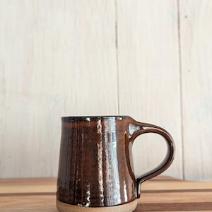 Puede incluir: Una taza de cer&aacute;mica marr&oacute;n con un esmalte moteado y un borde marr&oacute;n claro. La taza tiene un cuerpo redondeado y un asa.