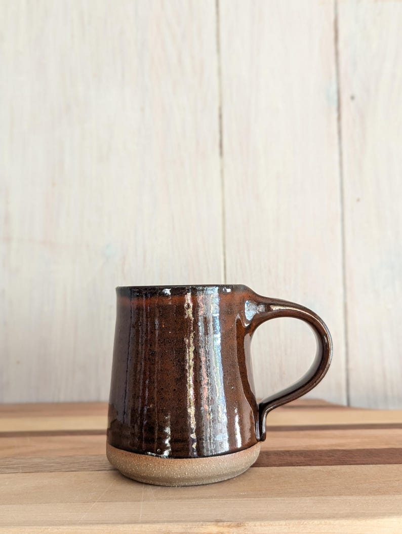 Puede incluir: Una taza de cer&aacute;mica marr&oacute;n con un esmalte moteado y un borde marr&oacute;n claro. La taza tiene un cuerpo redondeado y un asa.