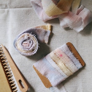 May include: A close-up of a wooden comb and a roll of colourful fabric ribbon. The ribbon is a mix of pink, purple, and yellow shades. The fabric is frayed and has a rustic look.