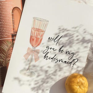 May include: Watercolor illustration of a champagne glass filled with pink liquid and a pink bow. The text "will you be my bridesmaid?" is written in elegant script. The card is on a white surface.