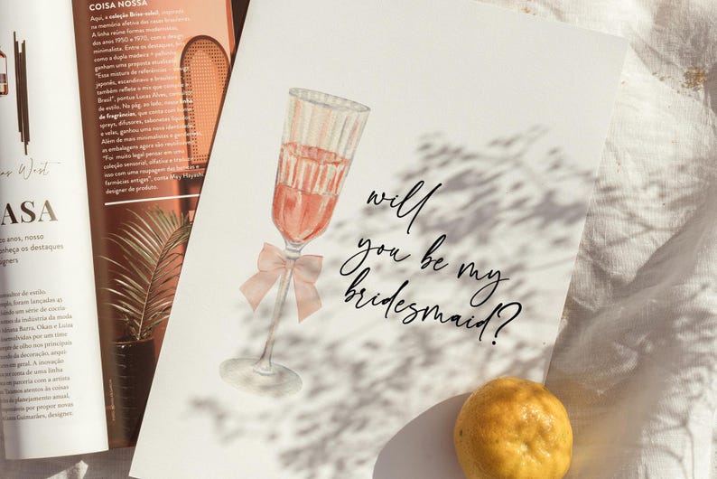 May include: Watercolor illustration of a champagne glass filled with pink liquid and a pink bow. The text "will you be my bridesmaid?" is written in elegant script. The card is on a white surface.