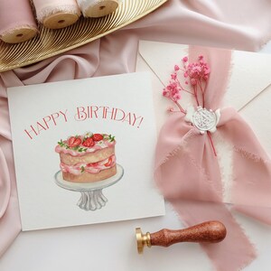 May include: A birthday card with a watercolor illustration of a cake with strawberries, the words "HAPPY BIRTHDAY!" in red, and a pink ribbon tied around an envelope. A wooden stamp is also present.