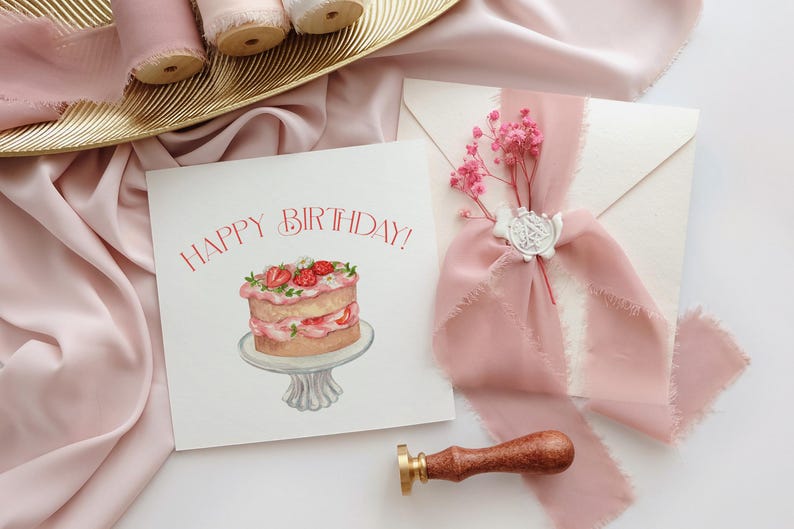May include: A birthday card with a watercolor illustration of a cake with strawberries, the words "HAPPY BIRTHDAY!" in red, and a pink ribbon tied around an envelope. A wooden stamp is also present.