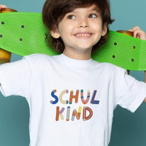 Könnte beinhalten: Ein weißes T-Shirt mit dem Text "SCHUL KIND" in bunten Buchstaben. Das Kind hält ein grünes Skateboard. Der Hintergrund ist einfarbig türkis.