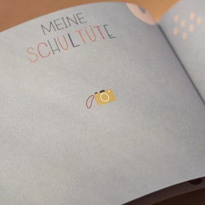 May include: A light blue book with the text "Meine Schult&uuml;te" in colorful letters. A small yellow camera with a red strap is on the page.