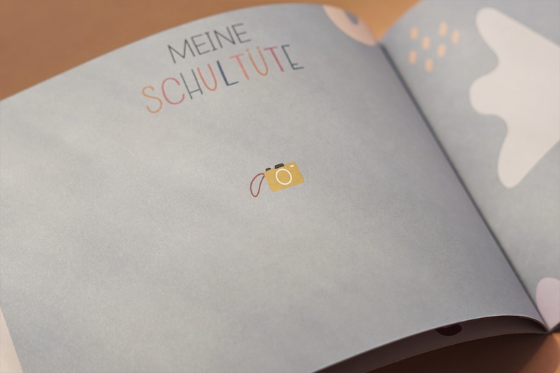 May include: A light blue book with the text "Meine Schult&uuml;te" in colorful letters. A small yellow camera with a red strap is on the page.