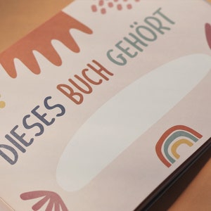 May include: A pink book with the text "DIESSES BUCH GEH&Ouml;RT" in blue and green lettering. The book has a white space for writing a name and is decorated with colorful abstract shapes.
