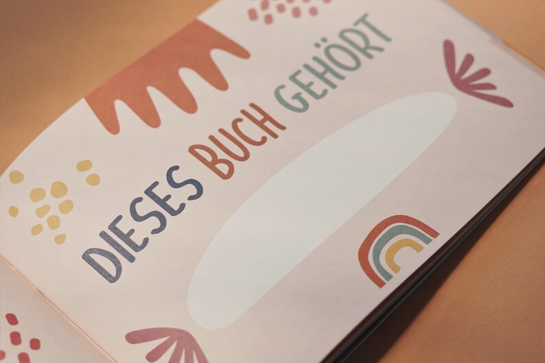 May include: A pink book with the text "DIESSES BUCH GEH&Ouml;RT" in blue and green lettering. The book has a white space for writing a name and is decorated with colorful abstract shapes.