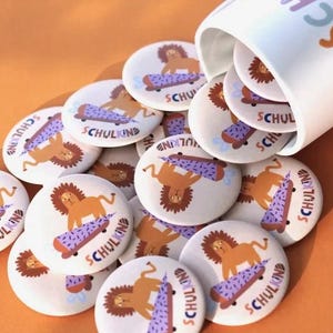 Könnte beinhalten: Weiße runde Buttons mit einem Cartoon-Löwen auf einem Skateboard. Das Skateboard ist lila mit einem rosa und braunen Design. Das Wort "SCHULKIND" ist in Braun auf die Buttons gedruckt. Ein weißer Becher schüttet die Buttons auf eine orangefarbene Oberfläche.