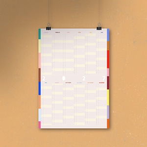 Könnte beinhalten: Ein Jahreskalender 2022 mit minimalistischem Design. Der Kalender ist auf hellbeigem Papier gedruckt und ist in Monate mit farbigen Registerkarten unterteilt. Der Kalender hängt an zwei schwarzen Büroklammern an einer hellbraunen Wand.