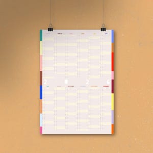 May include: A yearly calendar with a cream-colored background, hanging from black clips. The calendar has columns for each month and rows for each day. The left and right edges have colorful vertical stripes. The year "2026" is printed in the center.