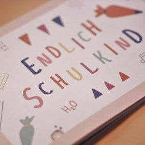 May include: A pink card with the text "ENDLICH SCHULKIND" in colorful letters. The card is decorated with colorful geometric shapes, including triangles, a protractor, and a musical note.