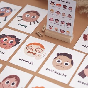 May include: A set of flashcards featuring illustrated faces with different emotions, each card has a German word describing the emotion. The words include "erstaunt", "mutig", "besorgt", "verletzt", "entt&auml;uscht", "ersch&ouml;pft", "verlegen".