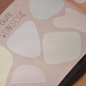 May include: A pink background with the text "GUTE W&Uuml;NSCHE" in blue and white lettering. There are five white, cream, and light pink abstract shapes on the background.